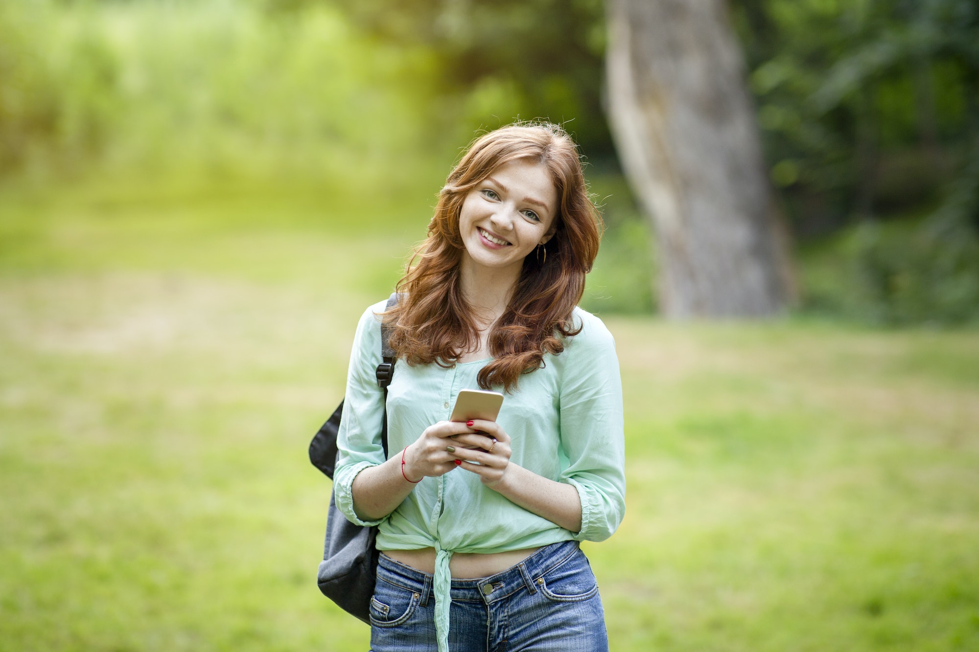 Potrait of cute redhead teen girl with backpack and smartphone outdoors
